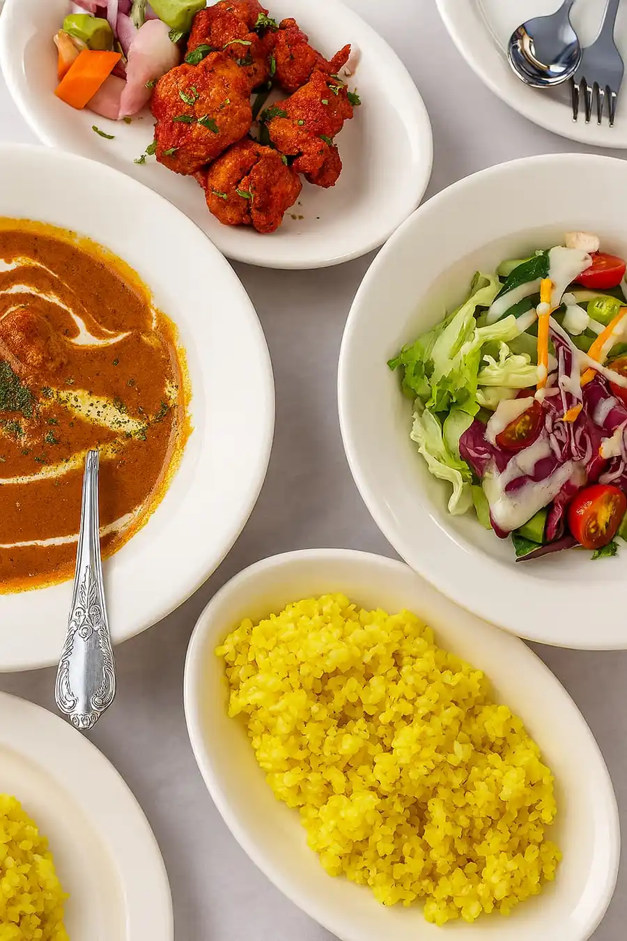 A colorful Indian lunch set featuring curry with cream swirl, yellow turmeric rice, fresh salad with dressing, and tandoori chicken served on white plates.